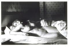 This photograph depicts a nude woman reclining on a bed, smiling and gazing back at the viewer. 