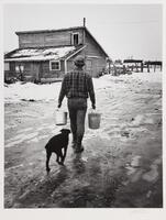 A back view of a man walking holding two metal buckets. There is a dark colored dog walking next to him.
