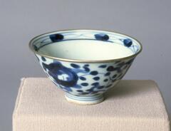 A petite cup with an abstract floral design in blue and white covering the outside of the cup and inside rim. The outside rim is gold and slightly everted. It has a large foot with blue stripes.