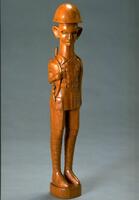 Carved wooden figure of a soldier. The general shape is cyclindrical and elongated on the vertical axis. The helmet, uniform, and gun are styled after WW2 era armaments.  The jacket, gun, and face are detailed.