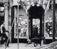 Blurry image of a man walking a dog on a leash past busy shops in the background.