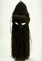 Mask is made entirely of blackish-brown dyed and molded raffia fiber; face has bulging forehead, deeo set narrow eyes, bulbous nose, and raffia “beard.” Top of head has cone-like crest of small fiber knots.<br />