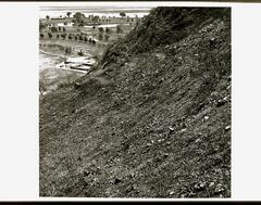 This is a black and white photograph showing a rocky, barren hillside above  a grassy golf course in the vally below. The rocky terrain on the slope is depicted in great detail. The golf course has a large pond and tree lined fairways.