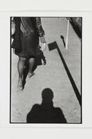 A woman seen from behind walks away from the camera on a sidewalk. A shadow of a person falls onto the concrete just behind her.