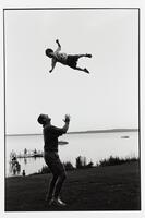 A man tossing a child in the air. They are on a grassy field with water in the background with a dock and other people.