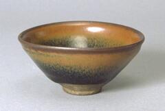 A deep, conical bowl on a straight foot ring with subtle rim articulation, covered in a thickly applied, dark, iron-rich black glaze with lighter russet-brown hare's fur markings (兔毫盏 <em>tuhao zhan</em>).  The thick glaze thins at the rim to a russet-brown color.