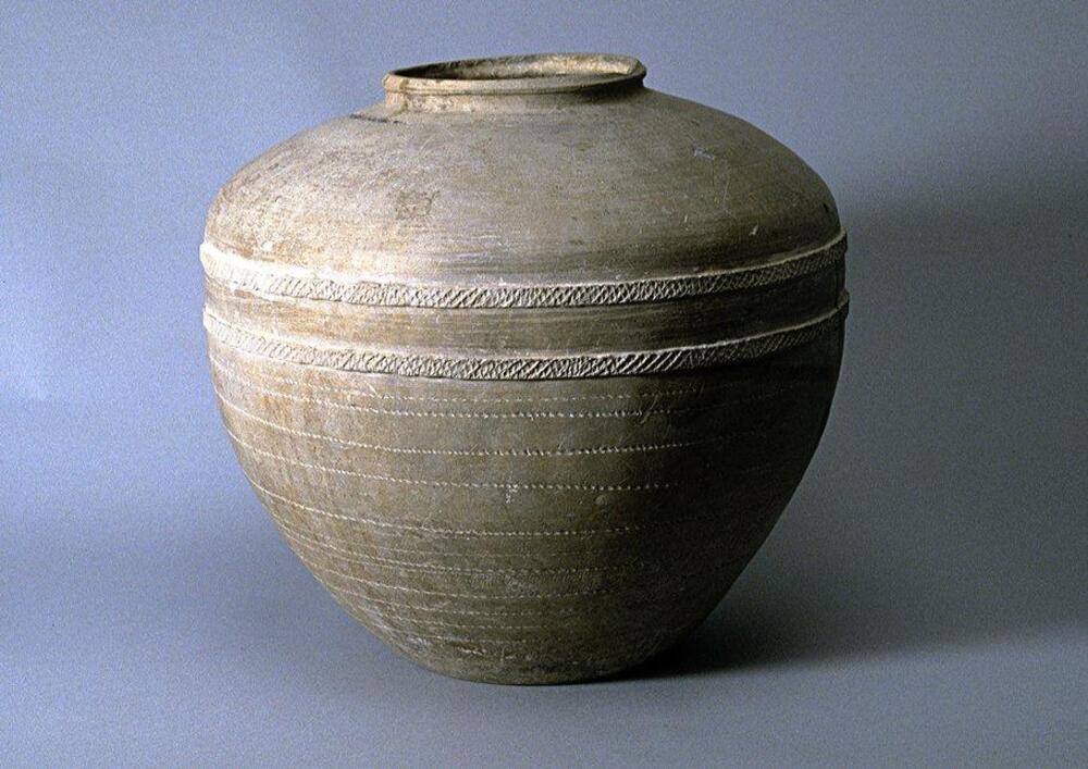 A light gray-buff earthenware <em>weng </em>甕 jar with a wide globular upper body, slightly angled shoulder and conical lower body on a flat base, and a short narrow neck with rim articulation. There are two appliqué coils around the upper body, with impressed rope decoration, and an impressed wheel or rope decoration around lower coiling on the lower portion of the body.