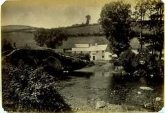 This photograph depicts a village with dwellings set in a hillside. A small bridge reaches over a river, which extends from the foreground through the middleground of the scene. 