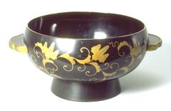 A rounded bowl with small stand underneath and two handles on either side. Gold covers the side of the handles and runs along the rim of the bowl. Wrapping around the outside of the bowl are gold floral patterns. Part of a bridal trousseau.