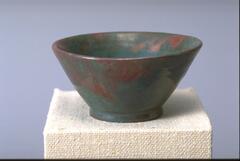 Bowl-shaped footed vessel covered with an iridescent glaze over a semi-matte glaze that creates the appearance of irregular patches of color ranging from a deep green-blue to a coppery-orange.