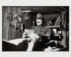 A mannequin in a military uniform holds a small book open with both hands. The walls and ceiling are made of unfinished wood planks. Photographs of posed people are pinned to the wall.