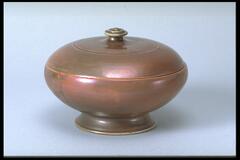 Footed ceramic bowl-shaped vessel with lid and knobbed handle covered in an iridescent copper glaze