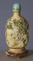 An ivory snuff bottle with design of green and yellow peonies and mandarin ducks. On the top of the snuff bottle is a mouthpiece with a blue stopper. The snuff bottle is sitting on a decorated wooden stand.