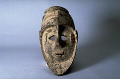 Carved wooden mask in the form of a oval-shaped face. The eyes are set close together with a small nose and mouth. The center of the face is sunken while the chin and forehead are raised. There are three small squares next to either eye and horizontal grooves on the forehead. 