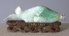 A Jadeite snuff bottle that is shaped like a fish. The body of the fish is carved with scales, the head has caring of eyes and the mouth of the fish is where the opening of the snuff outlet is with a cap at the very end of it. The back of the snuff bottle is shaped like the tail fin of the fish. The front (head) of the fish is green and fades into white at the body of the fish. It is sitting on a decorated wooden stand.