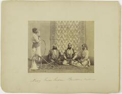 A black and white image of four men: one stands to the left, facing right, while the other three sit on their knees and hold swords in their hands.  A hookah stands to the left.
