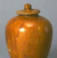 Stoneware jar with flaring foot and high rounded shoulders on a tapered body. It has an everted, flared foot, and short narrow neck covered with a flat lid with conical knob. The top three-quarters of the jar is covered in an amber-colored glaze. 