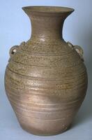 A brown stoneware, tall jar with a bulbous body and a tall flaring neck with direct everted rim, on a flat base. It has appliqué bands around the body and rim alternating with incised scroll work, combed wavy lines incised around neck and rim, and two lug handles with appliqué coil finials on opposing shoulders. The upper half is covered in a green ash glaze.