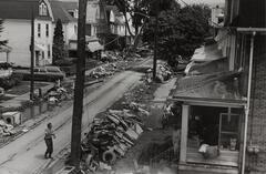 A man standing in the middle of a street that had been flooded. There are piles of debris in front of the houses.