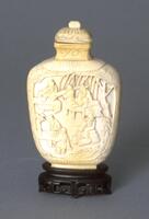 An ivory snuff bottle shaped like a flask. Carved in high relief are people sitting on chairs around tables with trees in the background. On the top of the flask is a stopper. The snuff bottle sitting on an incised wooden snuff bottle.