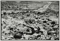 Image of a grassless field of mud and rock. A single pole and a set of vehicle tracks can be seen in the distance. In the foreground, the body of a man is tipped on his side, limbs stiff and bent at awkward angles.