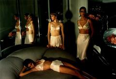 A color photograph of three models in an interior space, each wearing white satin lingerie. Two models stand in the background next to a large full-length wall mirror, in which their reflections repeat. In the foreground, a third model reclines on a large sofa.