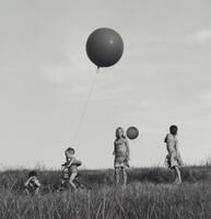 A photograph of four children standing in a field of grass. Two girls stand on the right side of the frame, one holding a balloon that blows in front of her. An infant on a bicycle stands to her left, holding a second balloon that has moved into the foreground, appearing larger due to its proximity to the lens.