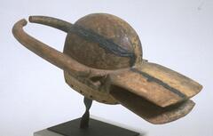 Wooden helmet-shaped mask with a large central dome. From the front two flat plates form a mouth, with triangular ears on each side while two curved horns extend back. There is a crack along the center of the mask that appears to have been repaired locally with pitch or tar.