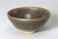 A conical bowl on a tall straight foot ring, covered in a dark brown-black glaze with brown oil spot markings ( 油滴釉 <em>you di you)</em>, thinning to brown at the rim. There are three chips to the rim.