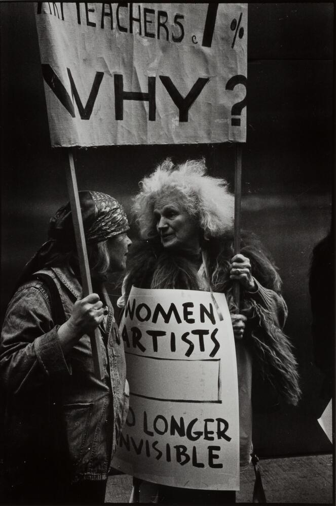 Two women holding signs at a protest march. They are talking together on the side of a street.