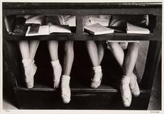 A photograph of the legs of four ballerinas, visible under a desk. They each wear ballet slippers and socks. The desk has an opening where books and wallets are stored for their lesson.