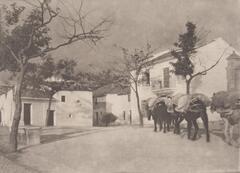 This image depicts two teams of horses, laden with goods on pack saddles as they walk through a square, between large white buildings. 