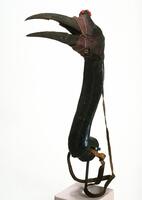 Carved wood headdress in the form of a bird's head and neck. The beak is open and the head is covered with leather, some of which is red. There is also string wrapped around the head of the bird. The crest above the beak is covered with red seeds, possibly abrus seeds. The long, curved neck is attached to a flat disc, through which leather straps have been inserted. The strap at the bottom of the neck is also attached to the back of the bird's head as well. 