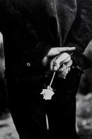 A black and white photo of a man holding his hands behind his back. In his hands is a white rose.