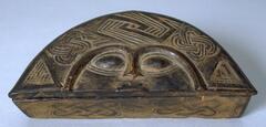 A wood box of semi-circular form, with a stylized form of a human face incised on the lid. The sides of the box, and around the face, are incised with geometric motifs, and two small holes are pierced in the lid and front of the box to fasten. The sides of the box are also covered with red powder. 