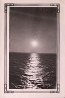 The horizon line of a body of water. The sun is a white spot in the center of the image, reflecting down on the surface of the water. The photograph is surrounded by a printed frame.