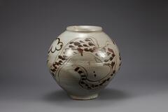 Round porcelain jar with iron pigment under colorless glaze. An abstract dragon spirals around and up the body of the piece, marked by quick brushstrokes indicating scales and unrestrained swirls indicating features such as its head and feet. A slight valley in the contour of the jar marks where two separately thrown pieces were joined together.<br />
The foot is rather small for the size of the body.<br />
<br />
This is a white porcelain jar decorated with iron-painted dragon, which wraps around the jar three times, displaying dynamic strokes of brush. The dragon&rsquo;s head is not rendered; its two eyes have been tersely painted instead. Jars with iron painted dragons, rendered in an abstract from, were produced in large quantities in the 17th century; many of them were produed in regional kilns. Despite slight damage to its rim, this jar is preserved as intact. The central part of its body clearly shows that this jar was created by joining separately produced upper and lower halves.<br />
[Korean Col
