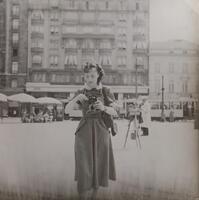 A woman in a shin-length dress with a white collar standing on a city street holding a camera. She is looking down and there are umbrellas and streetcars visible in the background.