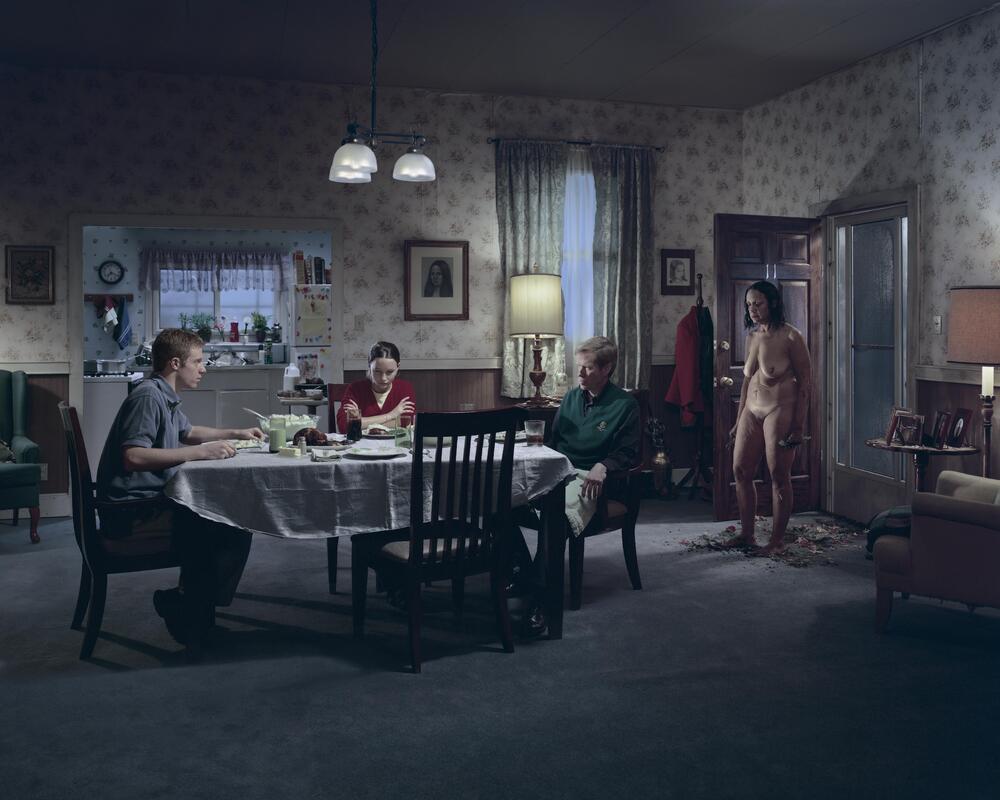 A color photograph of three people sitting around a dining room table. &nbsp;To the right, a nude woman stands inside, next to an open front door. &nbsp;There is dirt and trampled flowers at her feet.