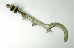 The wood handle of this sword is wrapped in metal wire and embellished with a design of two stacked discs. The cutting edge of the sword runs along the length of one side. The cutting edge is made up of three half circles with the last and largest half circle forming a large, pointed hook. 