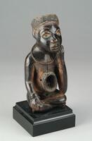 Carved wooden figure of a human. The umbilicus protrudes and is concave, which would have held magical/medicinal substances. The figure is posed with crossed legs and one hand supporting the head. The face is detailed, with glass eyes. A metal ring was placed around the neck of the figure. The top of the head is empty, but possibly contained magical substances as well. 