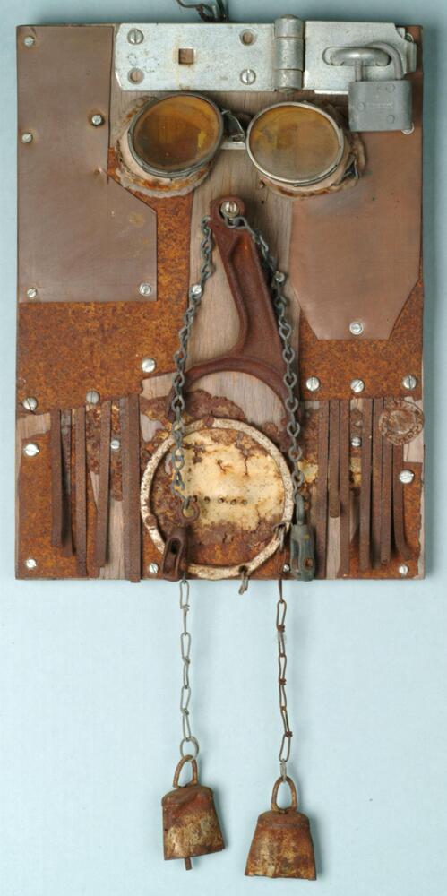 A mixed media assemblage consisting of welding goggles, rusted metal, a padlock, and two rusted bells hanging from a chain mounted on a wooden board with screws. The position of the objects, with goggles at the top, makes the assemblage look vaguely face-like.