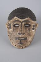 Oval shaped wooden mask with white pigment on the face and black pigment on the hair, which has a scalloped pattern near the forehead. There is also black pigment outlining the eyes, mouth, nose, as well as facial markings next to and below the eyes. Along the edge of the mask are small holes, perhaps to attach a now missing part of the costume. 