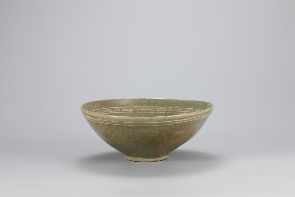 <p>This type of celadon was produced in large quantities during the 13th century when celadon with inlaid designs became more decorative. The upper part of the inner wall features a band of scroll design inlaid with white slip close to the rim, below which are four double concentric circles each containing a peony spray inlaid with black and white slip. Glaze was applied down to the rim of the foot. Three quartzite spur marks remain on the outer base. The entire inner surface features ne crazing. Two horizontal bands inlaid with white slip surround the upper part of the outer surface. The glaze was partially oxidized, tinged with brown.<br />
[<em>Korean Collection, University of Michigan Museum of Art </em>(2014) p.105]</p>
Shallow bowl with celadon glaze. Four peony designs encapsulated by a double-ringed circle float equally spaced along the inner curve of the bowl. A wavy fret design marks the inner rim, while the outer is marked by two or three incised lines.