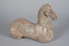 A red earthenware horse torso sculpture. Its muscular neck holds its narrow head high. The head is vividly sculpted, showing the musculature of the horse's face, flaring nostrils and an open mouth displaying the tongue. It has bulging eyes, ears pointing forward, and a trimmed mane. The body is stocky yet elegantly curved. There is loss to the legs. The horse is covered in white mineral pigment, with a blue flowing mane and cloud scroll on its rump, and red, white, and blue horse trappings and saddle. 