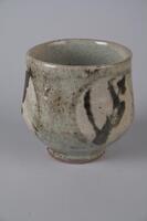 Stoneware tea bowl with short foot that flares out into the base of the tea bowl, and bends gently back inwards, until flaring slightly at the lip of the piece.  Underglaze design of a circle and stylized design or plant decorates the side of the bowl.