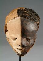 This bold and dramatic wooden Pende <em>mbangu</em> mask features a half-white, half-black face with a contorted visage.  The face has been carved in such a way that the features appearing on the left side, which has been pigmented black, are either positioned lower (the left ear and eye) or droop downwards (left sides of the nose and mouth). Moreover, the black portion of the face features several indentations representing scars. The resulting visual effect is one of facial asymmetry. A thick layer of white and black pigment has been applied to the face’s wooden surface, while black raffia fibers have been tightly woven onto a vegetable fiber cloth on the black half of the mask, completing the look.