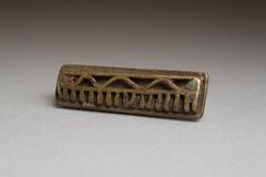Gold-weight in the shape of a rectangular base with a raised zig-zag pattern along one side and seventeen raised teeth along the other. 