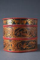 A round three-tiered wooden box with black, red, and gold lacquer. It has geometric and animal designs.