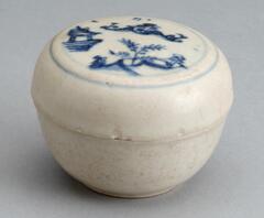 A stoneware ceramic box, with a rounded bowl and a nearly flat lid, decorated with a free-hand drawn landscape in underglaze cobalt blue on the lid, and then coated in a whitish glaze overall.